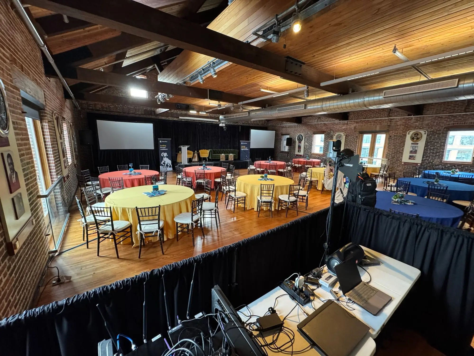 Front-of-house position at a corporate program in a historic brick hall: console, wireless rack, and laptop playback behind drape