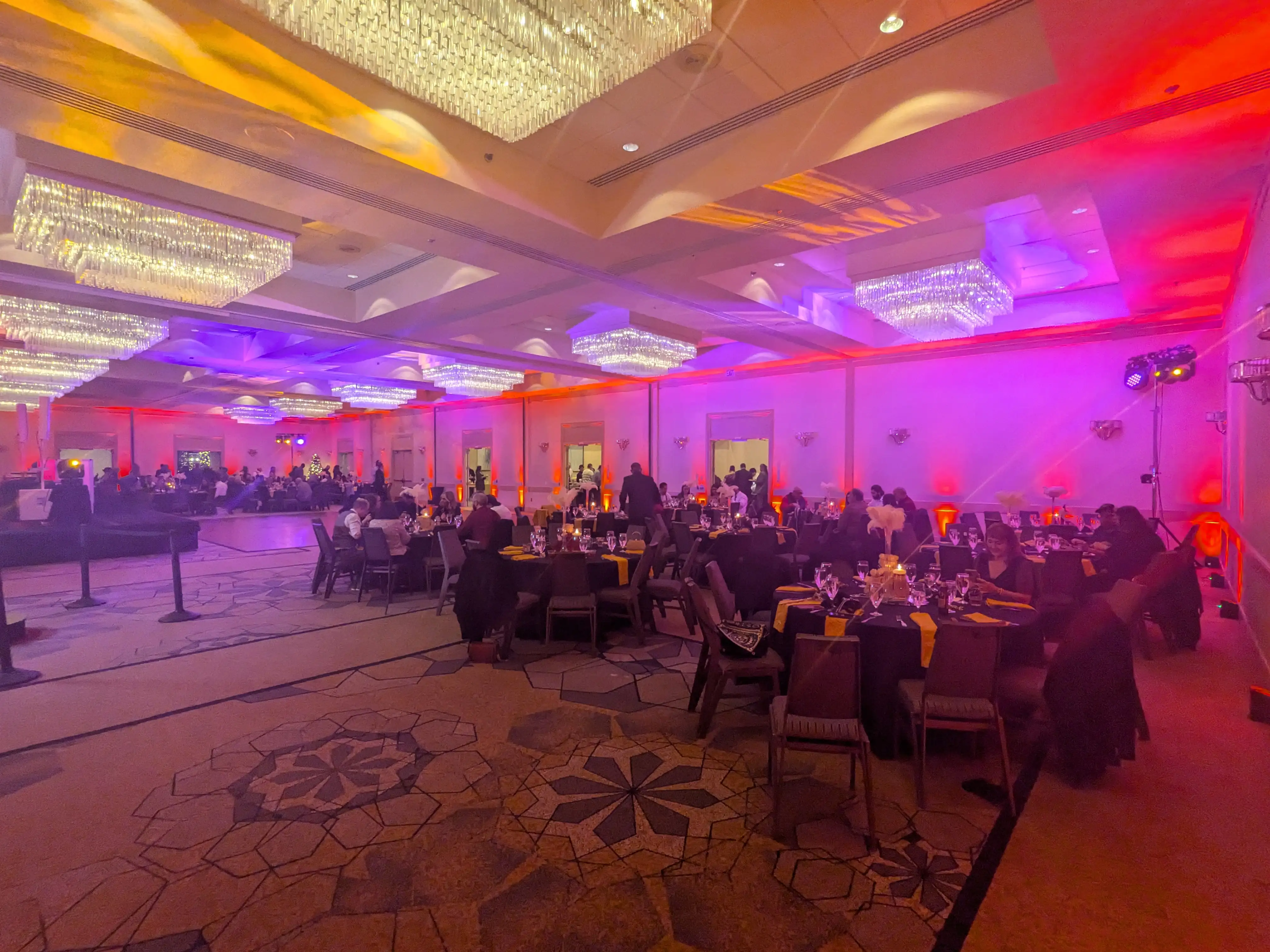 Corporate holiday party in a hotel ballroom washed in amber and violet uplighting, with a moving-head lighting fixture rigged house-right