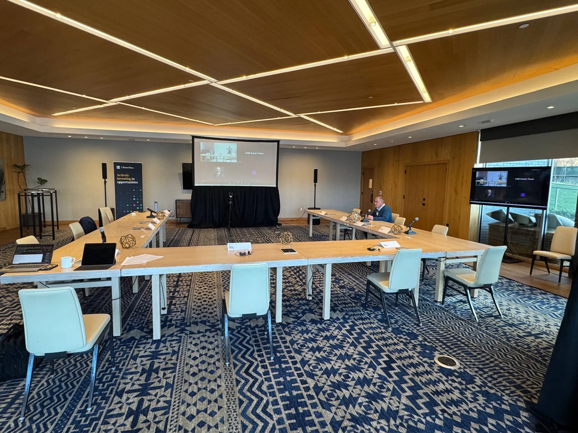 Boardroom set for a hybrid meeting — remote participants on the projection screen, tabletop microphones at every seat, a camera on tripod, and speakers flanking the screen
