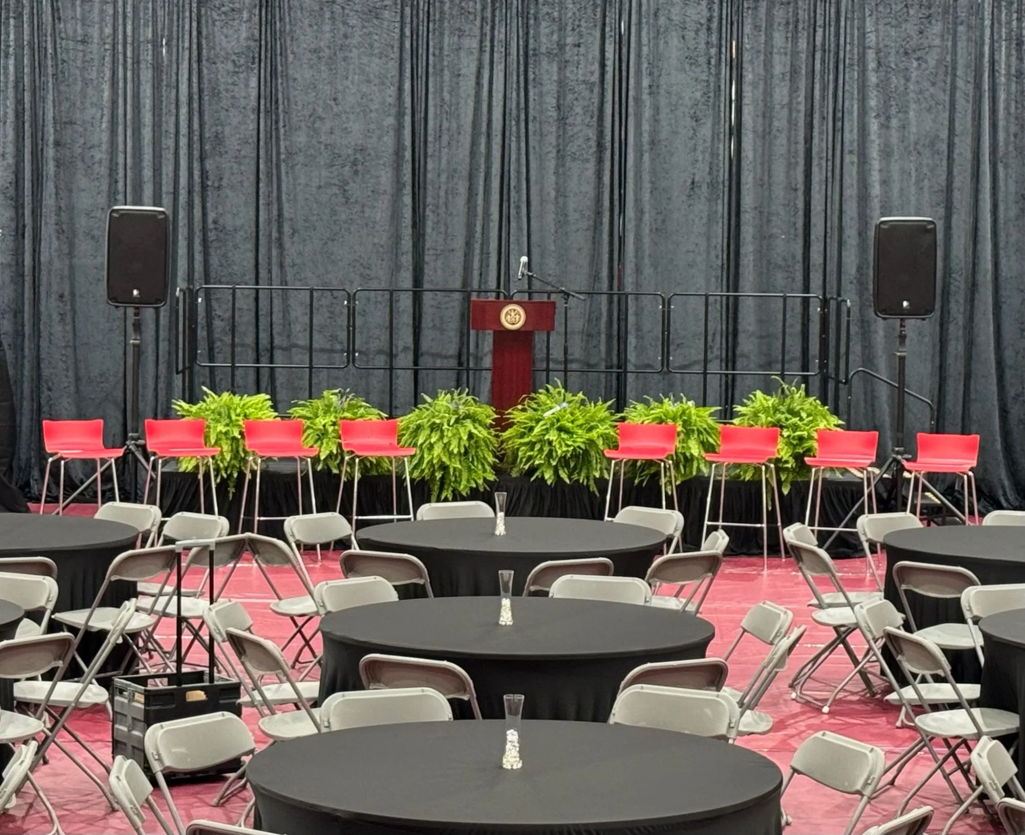 Skirted stage with a wooden podium, red panel seating, and ferns in front of a full gray drape wall, PA speakers on stands at each side